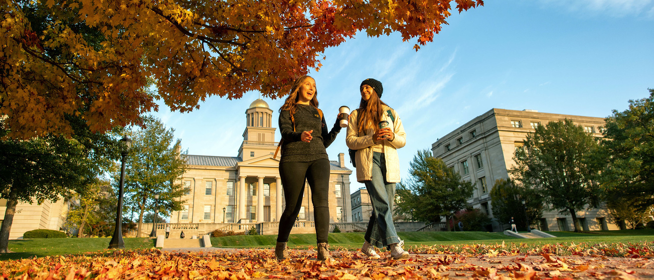 Two students walking down a leaf-covered sidewalk on the Pentacrest.